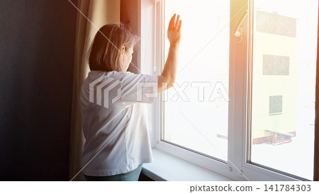 Senior woman standing at a bright window with her hand raised, engaging in social interaction from a distance, focusing on hope and connection during a time of loneliness Senior woman standing at a bright window with her hand raised, engaging in social interaction from a distance, focusing on hope and connection during a time of loneliness 141784303