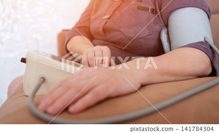 Senior woman sitting on a sofa, routinely checking her blood pressure with a home digital monitor and arm cuff, focusing on personal health and preventive care Senior woman sitting on a sofa, routinely checking her blood pressure with a home digital monitor and arm cuff, focusing on personal health and preventive care 141784304