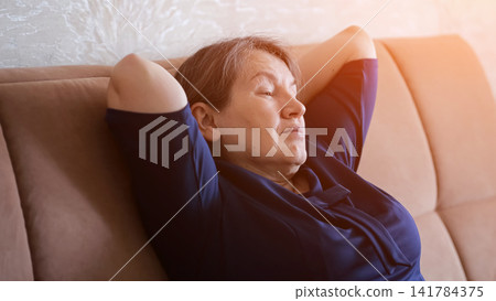 Elderly woman with closed eyes resting peacefully on a cozy sofa at home, enjoying calm relaxation and comfort in a serene indoor atmosphere with soft warm light Elderly woman with closed eyes resting peacefully on a cozy sofa at home, enjoying calm relaxation and comfort in a serene indoor atmosphere with soft warm light 141784375