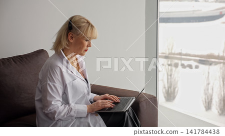 Blonde woman typing on a laptop, engaging in remote work or online education while sitting comfortably on a couch by a window with a winter landscape outside Blonde woman typing on a laptop, engaging in remote work or online education while sitting comfortably on a couch by a window with a winter landscape outside 141784438