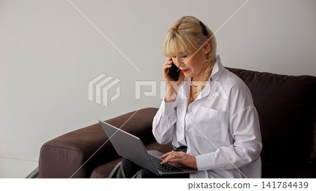 Woman engaging in remote work, managing business tasks from a home office setup, speaking on a mobile phone while simultaneously typing on a laptop, demonstrating efficiency and professionalism Woman engaging in remote work, managing business tasks from a home office setup, speaking on a mobile phone while simultaneously typing on a laptop, demonstrating efficiency and professionalism 141784439