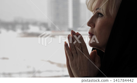 Senior woman in a black veil praying with clasped hands at a window, side profile showing quiet devotion, reflection and hope amid grief and solemn contemplation Senior woman in a black veil praying with clasped hands at a window, side profile showing quiet devotion, reflection and hope amid grief and solemn contemplation 141784442