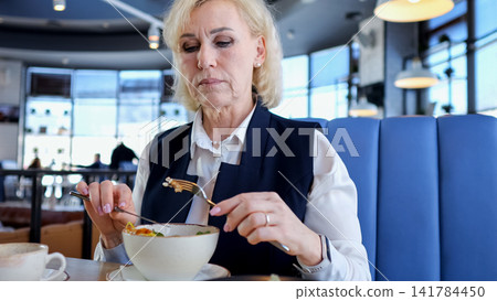 Caucasian Elderly Businesswoman Tasting Soup With Spoon In Quiet Cafe Booth, Thoughtful Expression, Warm Bowl, Focused On Flavors While Reviewing Documents On Table, Calm Reflective Mood. Caucasian Elderly Businesswoman Tasting Soup With Spoon In Quiet Cafe Booth, Thoughtful Expression, Warm Bowl, Focused On Flavors While Reviewing Documents On Table, Calm Reflective Mood. 141784450