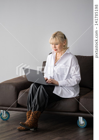 Mature blonde woman sitting comfortably on a brown couch, focused on typing on her laptop, embodying the modern freelance lifestyle and flexible remote work concept Mature blonde woman sitting comfortably on a brown couch, focused on typing on her laptop, embodying the modern freelance lifestyle and flexible remote work concept 141785198