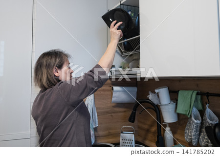 Woman reaching up to place a black pot lid in an open kitchen cabinet while cleaning the sink area in a modern home kitchen with white cabinets, wood backsplash, black faucet, hanging towels Woman reaching up to place a black pot lid in an open kitchen cabinet while cleaning the sink area in a modern home kitchen with white cabinets, wood backsplash, black faucet, hanging towels 141785202