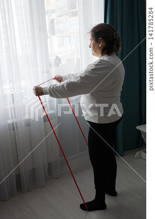 Senior woman using a red resistance band by a window for home fitness and rehabilitation, promoting strength, flexibility, independence, and active aging in a comfortable living room Senior woman using a red resistance band by a window for home fitness and rehabilitation, promoting strength, flexibility, independence, and active aging in a comfortable living room 141785284