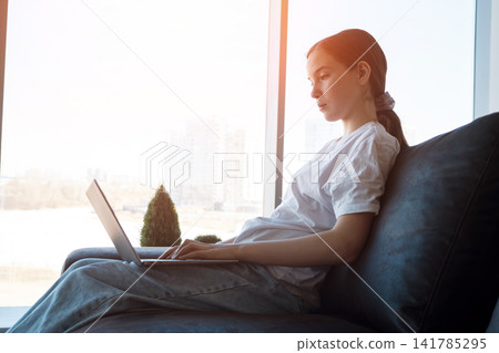 Young woman working remotely on laptop at window home Young woman working remotely on laptop at window home 141785295