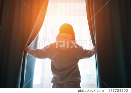 Person standing with back to camera, opening curtains at a window, letting bright morning light flood into the room, symbolizing a fresh start and hope Person standing with back to camera, opening curtains at a window, letting bright morning light flood into the room, symbolizing a fresh start and hope 141785300