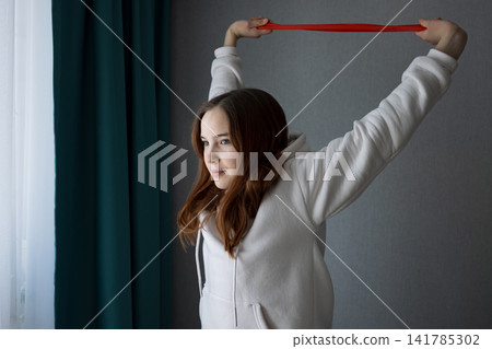 Young woman in a white hoodie performing resistance band exercises, stretching her arms above her head, focusing on fitness and wellness in a comfortable home environment Young woman in a white hoodie performing resistance band exercises, stretching her arms above her head, focusing on fitness and wellness in a comfortable home environment 141785302