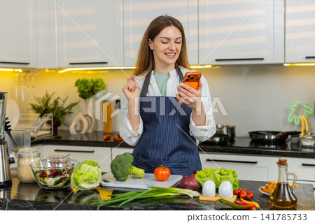Middle-aged woman smiling at smartphone in home kitchen celebrating winning message with good news Middle-aged woman smiling at smartphone in home kitchen celebrating winning message with good news 141785733