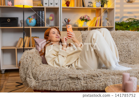 Young woman on home sofa smiling at smartphone screen while relaxing on couch and scrolling online Young woman on home sofa smiling at smartphone screen while relaxing on couch and scrolling online 141785752