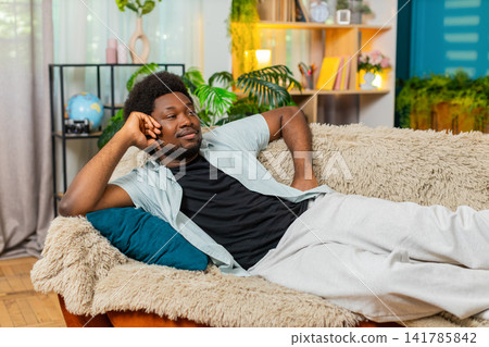 African American young man on home sofa settling with pillow and lying down for calm rest indoors African American young man on home sofa settling with pillow and lying down for calm rest indoors 141785842