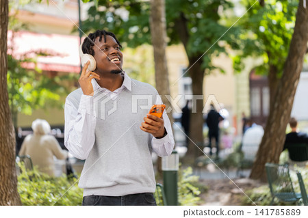 African American young man listening to music walking in city street with headphones and smartphone African American young man listening to music walking in city street with headphones and smartphone 141785889
