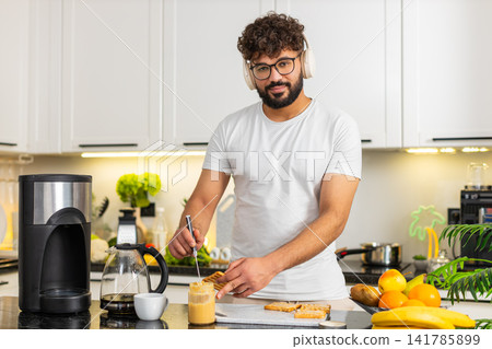 Arabian young man listening music in headphones making toast with peanut butter breakfast in kitchen Arabian young man listening music in headphones making toast with peanut butter breakfast in kitchen 141785899