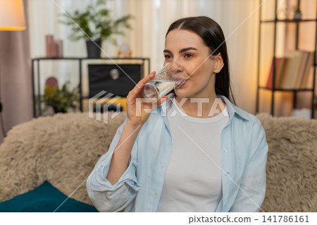 Young woman sitting on sofa drinking filtered water feeling thirsty advertising healthy good habit Young woman sitting on sofa drinking filtered water feeling thirsty advertising healthy good habit 141786161