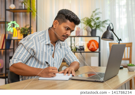 Indian man on remote learning uses laptop and notebook writes notes completes test quiz exam online Indian man on remote learning uses laptop and notebook writes notes completes test quiz exam online 141786234