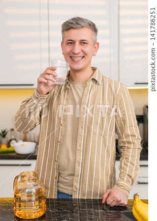 Middle-aged man drinking water in kitchen at home with smile after diet meal for weight loss routine Middle-aged man drinking water in kitchen at home with smile after diet meal for weight loss routine 141786251