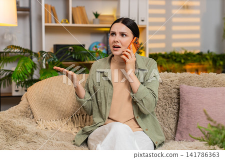 Young woman on home sofa arguing on smartphone expressing anger during emotional phone call indoors 141786363
