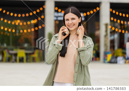 Young woman removing wireless headphones walking in city street smiling closeup portrait outdoors Young woman removing wireless headphones walking in city street smiling closeup portrait outdoors 141786383