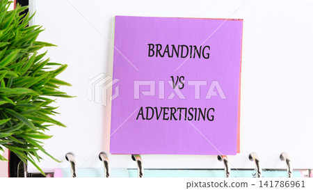 A note with the words branding and advertising is attached to a clipboard in an office space. A small green plant is also visible in the scene A note with the words branding and advertising is attached to a clipboard in an office space. A small green plant is also visible in the scene 141786961