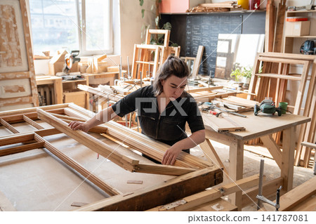 Confident woman in carpentry workshop works at workbench with vintage wooden window frame. Confident woman in carpentry workshop works at workbench with vintage wooden window frame. 141787081