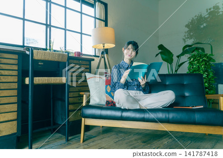 一位面帶微笑的年輕日本女子坐在沙發上,打開著筆記本。 一位面帶微笑的年輕日本女子坐在沙發上,打開著筆記本。 141787418
