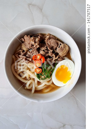 A bowl of udon noodles in a savory broth, topped with green onions, tempura, and a soft boiled egg. The thick, chewy noodles and rich flavors make it a comforting Japanese classic. A bowl of udon noodles in a savory broth, topped with green onions, tempura, and a soft boiled egg. The thick, chewy noodles and rich flavors make it a comforting Japanese classic. 141787517