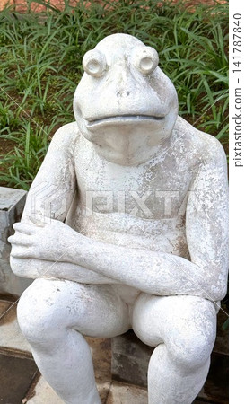 Weathered stone frog statue seated with crossed arms on garden bench, textured white surface showing worn paint, flaking details, and aged patina beside leafy groundcover Weathered stone frog statue seated with crossed arms on garden bench, textured white surface showing worn paint, flaking details, and aged patina beside leafy groundcover 141787840
