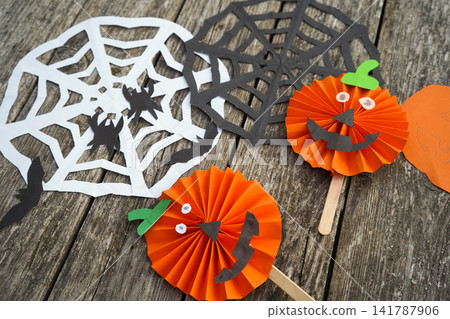 DIY Halloween pumpkin props made of pleated orange paper laid on a rustic wooden table near cutout spiderwebs DIY Halloween pumpkin props made of pleated orange paper laid on a rustic wooden table near cutout spiderwebs 141787906
