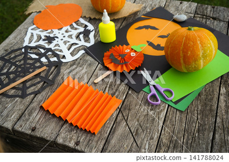 Spooky holiday craft mockup with handmade pleated paper pumpkin, paper cutouts and mini pumpkins on an old wooden surface Spooky holiday craft mockup with handmade pleated paper pumpkin, paper cutouts and mini pumpkins on an old wooden surface 141788024