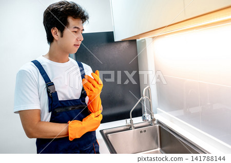 Asian male housekeeper putting on orange rubber gloves preparing himself for cleaning kitchen Asian male housekeeper putting on orange rubber gloves preparing himself for cleaning kitchen 141788144
