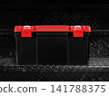 A black and red plastic storage box is placed on a black metal plate. 141788375