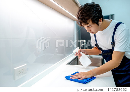 Asian male housekeeper holding spray bottle while wiping microfiber cloth on white countertop Asian male housekeeper holding spray bottle while wiping microfiber cloth on white countertop 141788662