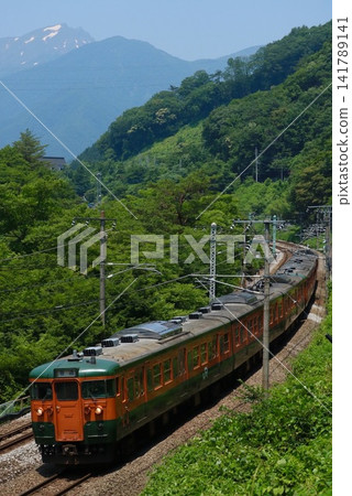 A 115 series Shonan-colored train running through the lush greenery with Mount Tanigawa in the background. A 115 series Shonan-colored train running through the lush greenery with Mount Tanigawa in the background. 141789141