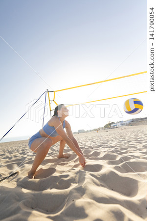 Woman plays volleyball on sandy beach during sunny day while bending down to hit the ball Woman plays volleyball on sandy beach during sunny day while bending down to hit the ball 141790054