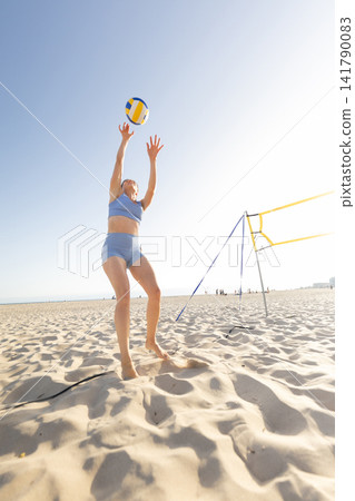 Woman plays beach volleyball on sandy shore during sunny day with ocean in background Woman plays beach volleyball on sandy shore during sunny day with ocean in background 141790083
