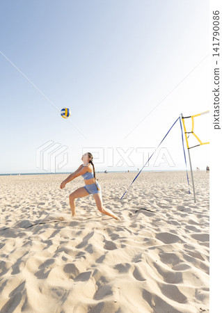 Girl plays beach volleyball on a sunny day with blue sky and sand in background Girl plays beach volleyball on a sunny day with blue sky and sand in background 141790086
