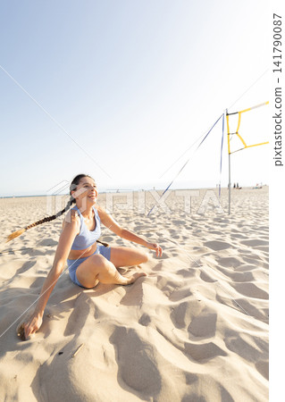 Person enjoys time on the beach while sitting in soft sand near a volleyball net under a clear blue sky 141790087