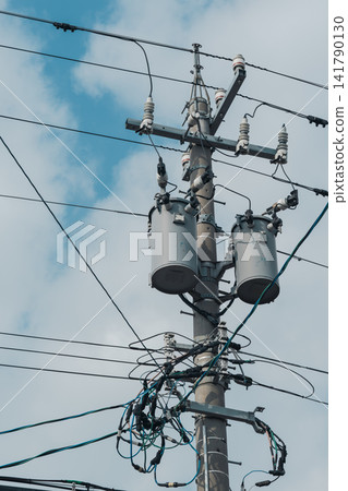 A utility pole with a transformer and a complex network of power lines stand against a blue sky. A utility pole with a transformer and a complex network of power lines stand against a blue sky. 141790130