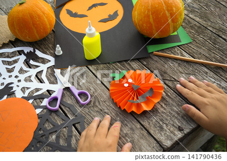 Over the shoulder view of a child carefully assembling parts of a paper pumpkin on a wooden stick Over the shoulder view of a child carefully assembling parts of a paper pumpkin on a wooden stick 141790436