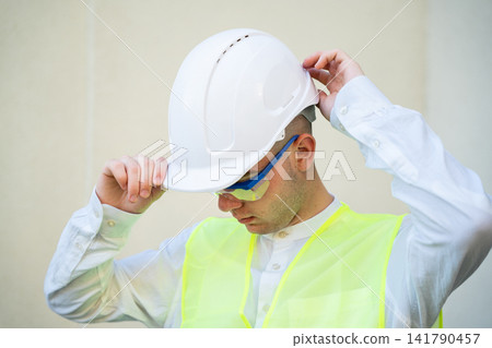 Worker adjusting hard hat while preparing for work in safety gear. Personal protective equipment and safety training in a construction environment 141790457