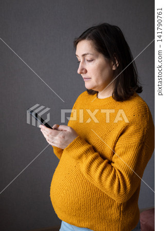 Pregnant woman in mustard sweater using a smartphone in a studio with gray background Pregnant woman in mustard sweater using a smartphone in a studio with gray background 141790761