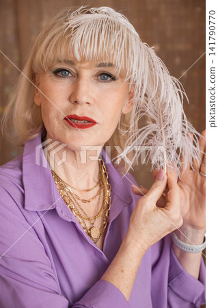 Elegant senior woman with blonde hair and red lipstick, wearing a purple shirt and gold necklaces, gently touching a white feather, portraying graceful aging and classic style Elegant senior woman with blonde hair and red lipstick, wearing a purple shirt and gold necklaces, gently touching a white feather, portraying graceful aging and classic style 141790770