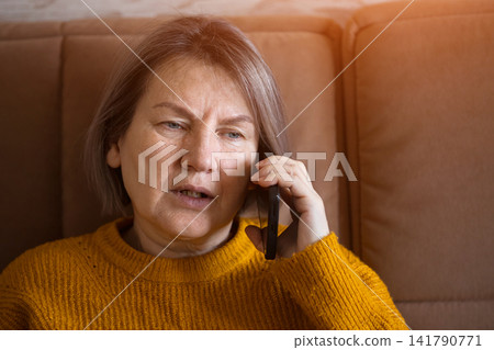 Concerned senior woman talking on mobile phone while sitting on a sofa at home Concerned senior woman talking on mobile phone while sitting on a sofa at home 141790771