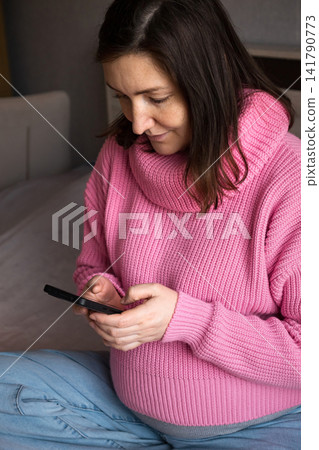 Pregnant woman in pink sweater using a smartphone while sitting on a sofa at home Pregnant woman in pink sweater using a smartphone while sitting on a sofa at home 141790773