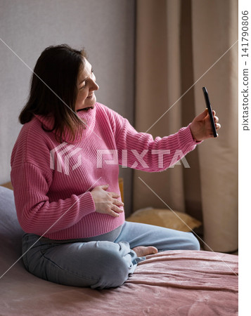 Pregnant woman in pink sweater holding her belly and using a smartphone while sitting on a bed Pregnant woman in pink sweater holding her belly and using a smartphone while sitting on a bed 141790806