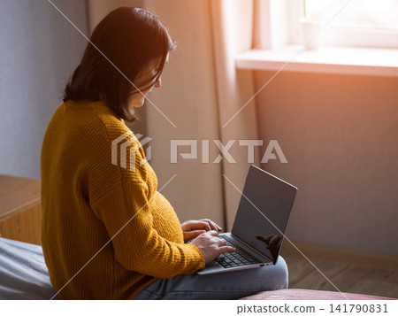 Pregnant woman using a laptop while sitting on a bed in a bright home bedroom Pregnant woman using a laptop while sitting on a bed in a bright home bedroom 141790831
