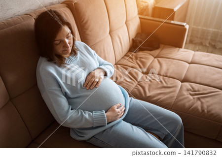 Pregnant woman resting on a beige sofa in a warm home living room 141790832