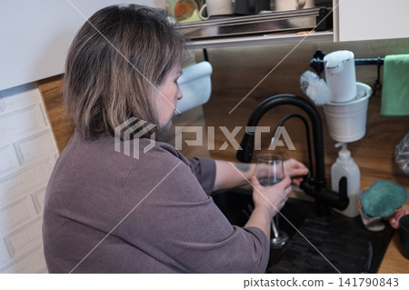 Woman washing a glass at a kitchen sink in a modern home interior with open shelving and cleaning items, black faucet, wood backsplash, white cabinets, hanging towels, and soft natural light Woman washing a glass at a kitchen sink in a modern home interior with open shelving and cleaning items, black faucet, wood backsplash, white cabinets, hanging towels, and soft natural light 141790843