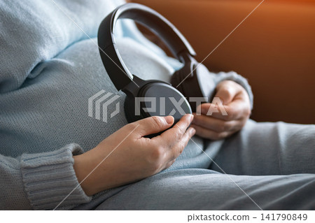 Pregnant woman relaxing on a sofa with headphones and listening to music at home Pregnant woman relaxing on a sofa with headphones and listening to music at home 141790849